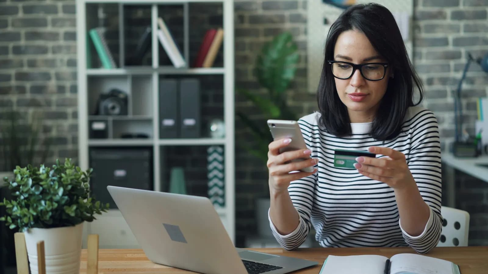 Mujer sentada en un escritorio con la notebook abierta, mirando el celular mientras sostiene una tarjeta de crédito