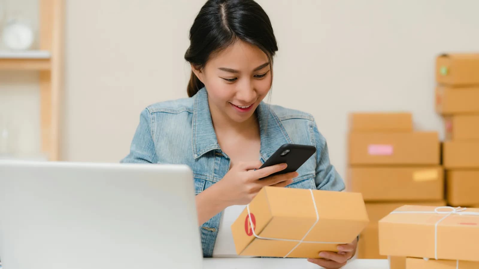 Mujer en su escritorio de casa revisando con el celular una caja de cartón, con la notebook abierta y otras cajas listas para enviar