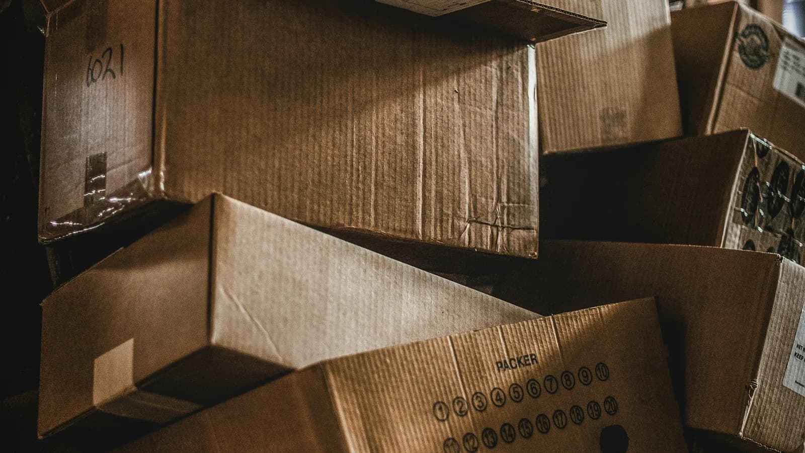 Cajas de cartón marrón apiladas y desordenadas en un depósito con poca luz, una con casilleros numerados impresos